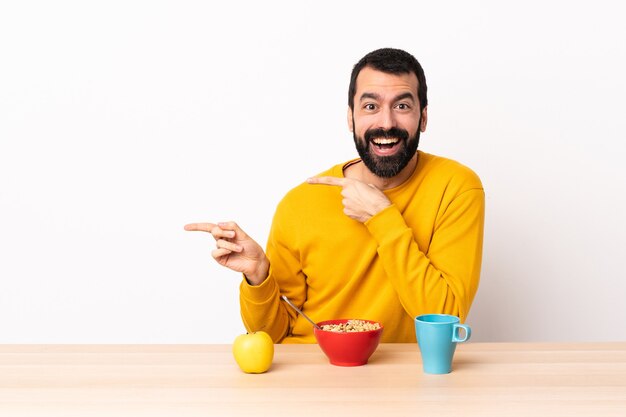 Homem caucasiano tomando café da manhã em uma mesa surpreso e apontando o lado.