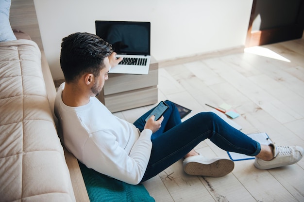 Homem caucasiano usando seu laptop enquanto está sentado no chão e enviando mensagens de texto - pandemia de trabalho remoto