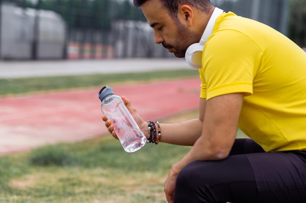 Homem chateado com fones de ouvido sem fio olhando para garrafa de água fresca sentado após treino em esportes
