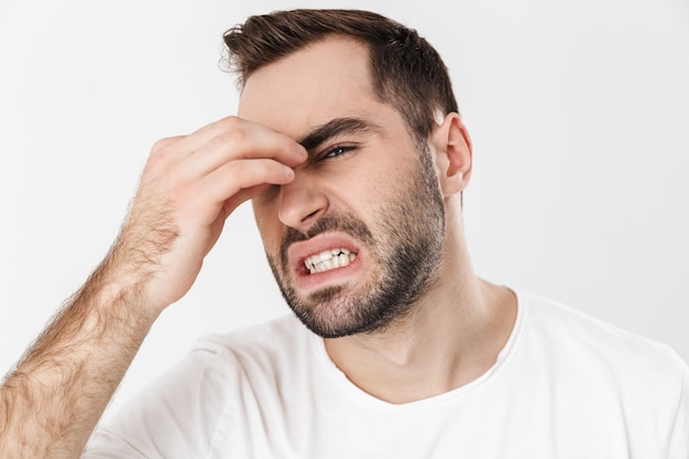 Homem chateado, vestindo uma camiseta em branco, em pé, isolado sobre uma parede branca, sofrendo de uma forte enxaqueca