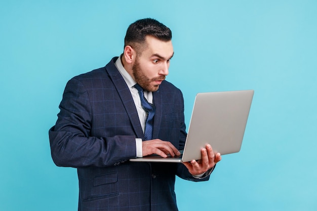 Homem chocado com barba vestindo terno estilo oficial, olhando para a tela do laptop com a boca aberta e olhos grandes surpresos com erro do sistema de operação tiro de estúdio interno isolado em fundo azul