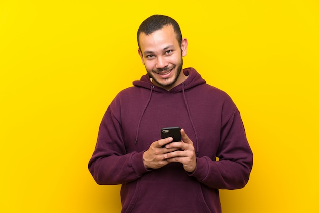 Homem colombiano com moletom sobre parede amarela, enviando uma mensagem com o celular