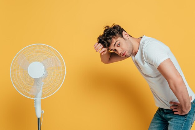 Homem com a mão no quadril olhando para a câmera e inclinando-se perto do ventilador elétrico em amarelo