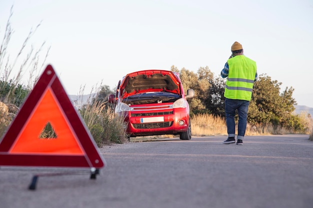 Homem com atitude relaxada encalhado na estrada ao lado do carro quebrado falando ao telefone