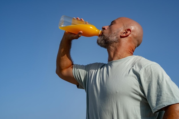 Foto homem com barba bebe suco de laranja recém-espreitado de uma garrafa enquanto está de pé contra o fundo do mar