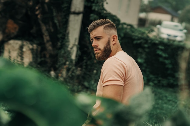 Homem com barba e camisa rosa posando no parque verde.