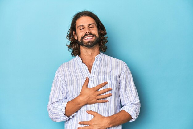 Homem com barba em camisa listrada azul estúdio azul ri feliz e se diverte mantendo as mãos no estômago