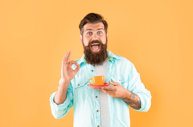 Homem com barba espantado na camisa com café mostra gesto okey no café de fundo amarelo