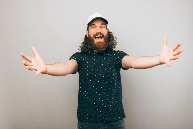 Homem com barba está sorrindo para a câmera enquanto quer dar um abraço sobre uma parede cinza