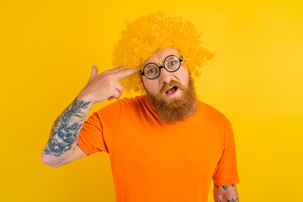 Foto homem com barba, peruca amarela e óculos fazendo um gesto de arma com a mão
