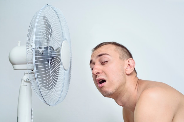 Homem com barba por fazer sofre com o calor e tentando se refrescar perto do ventilador.