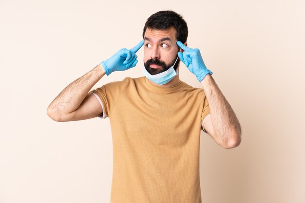 Homem com barba, protegendo do coronavírus com uma máscara e luvas sobre parede isolada, tendo dúvidas e pensando