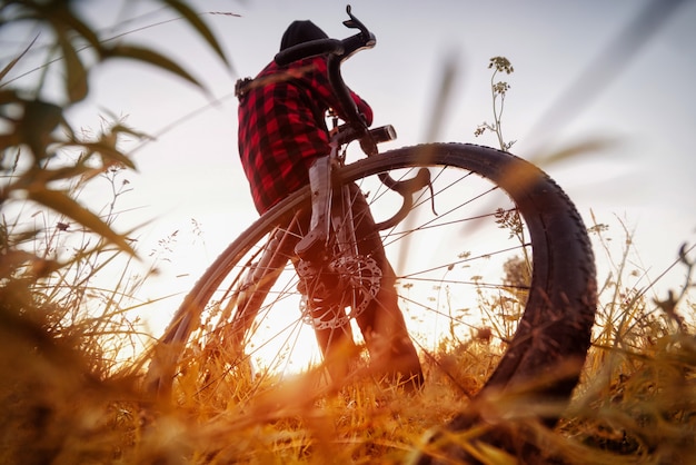 Homem com bicicleta no campo. visão de grande angular de um ciclista sentado em sua bicicleta ao nascer do sol em um campo com grama. conceito de estilo de vida ativo.