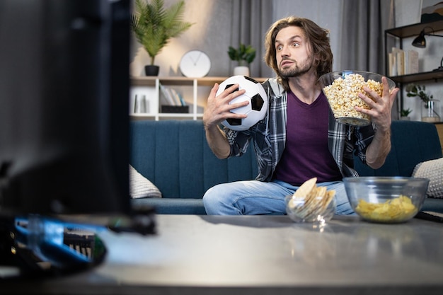 Homem com bola nas mãos sentado no sofá e assistindo futebol