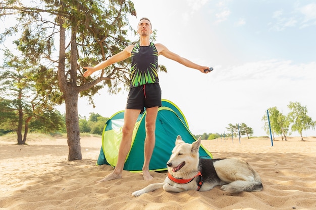 Homem com cachorro perto da barraca sob uma árvore em uma praia arenosa