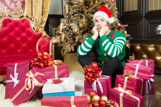 Homem com caixas de presentes na árvore de Natal. Homem prepara pacotes de presentes na véspera de ano novo. Homem fantasiado de Papai Noel comemora o ano novo. O Papai Noel para aqui.