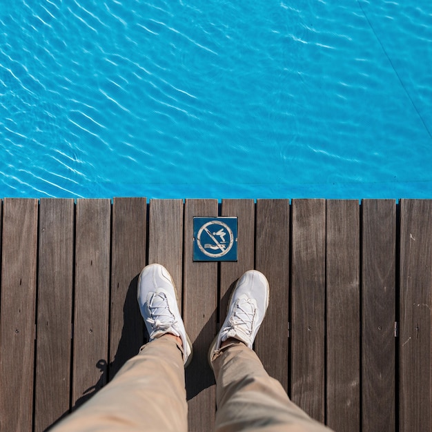 Homem com calça bege e tênis branco da moda fica em um piso de madeira perto de uma piscina com água azul não mergulhe sinal vintage