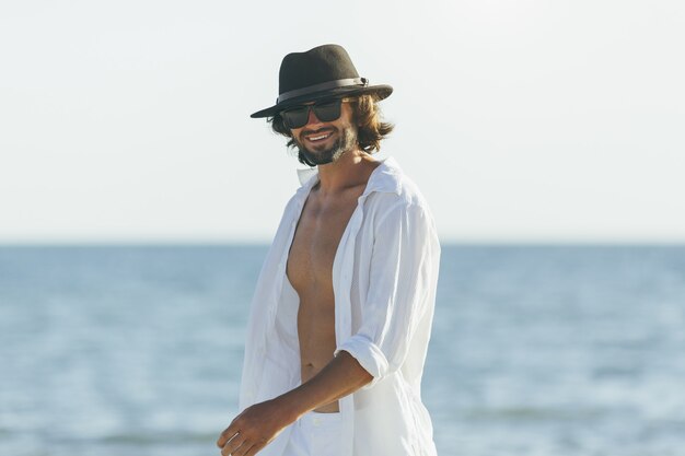 Homem com camisa branca, chapéu preto e óculos de sol andando feliz na praia