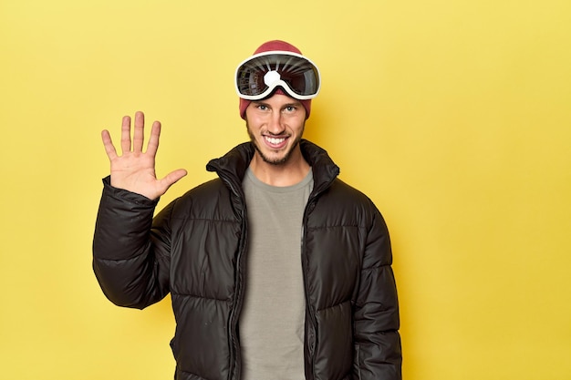 Homem com casaco de inverno com óculos de neve amarelo sorrindo alegre mostrando o número cinco com os dedos