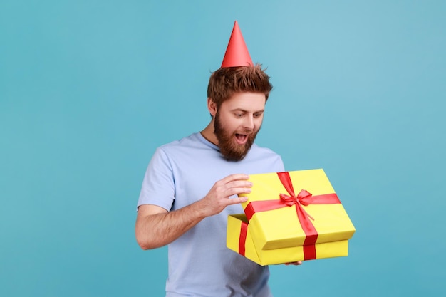 Homem com chapéu de cone de festa segurando a caixa de presente aberta e olhando para o presente com um olhar animado desembalando o presente desfrutando de uma incrível surpresa de aniversário Foto de estúdio interior isolada em fundo azul