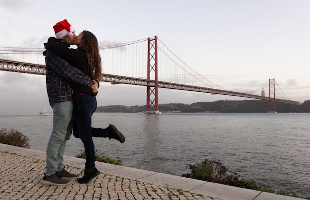 Homem com chapéu de Natal e mulher a beijar-se na ponte 25 de Abril em Lisboa Conceito de turismo em Portugal