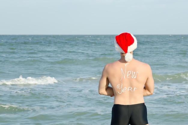 Homem com chapéu de Papai Noel com a inscrição "Ano novo" nas costas, em pé na praia