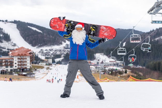 Homem com chapéu de papai noel com uma prancha de snowboard em uma estação de esqui.