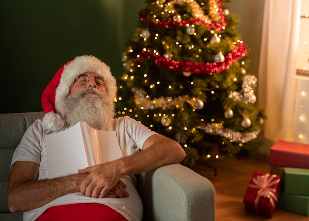 Homem com chapéu de papai noel dormindo no sofá em casa enquanto segura um livro