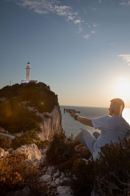 Homem com drone no penhasco perto do pôr do sol do mar acima do farol