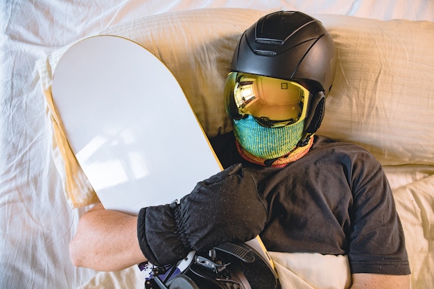 Homem com equipamento de esqui dormindo na cama com snowboard sonhando com montanhas