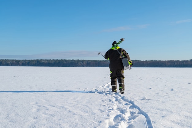 Homem com equipamento de pesca, caminhando em um lago congelado para longe em um dia ensolarado de inverno.