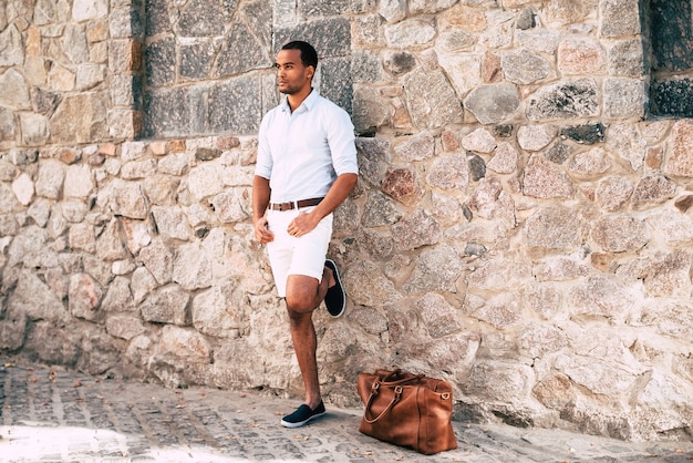 Homem com estilo. Comprimento total de um jovem africano bonito em roupas elegantes casuais em pé contra a parede de pedra ao ar livre com sua bolsa perto dele