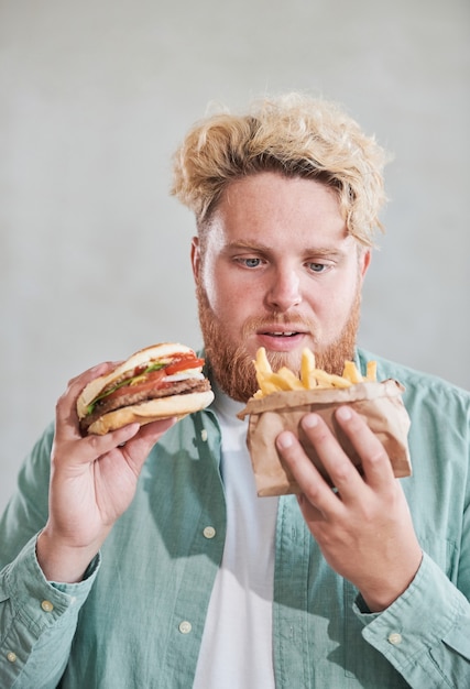 Homem com excesso de peso comendo batata frita e hambúrguer contra o fundo branco