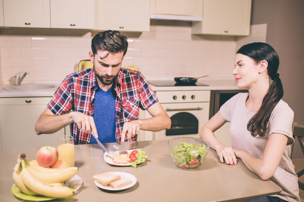 Homem com fome senta-se e mesa e corta a refeição em seu prato com garfo e faca.