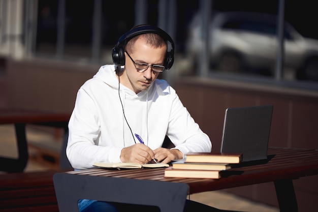 Homem com fones de ouvido está trabalhando em um laptop e escrevendo em um caderno sentado em uma mesa na rua. Distanciamento social durante o coronavírus
