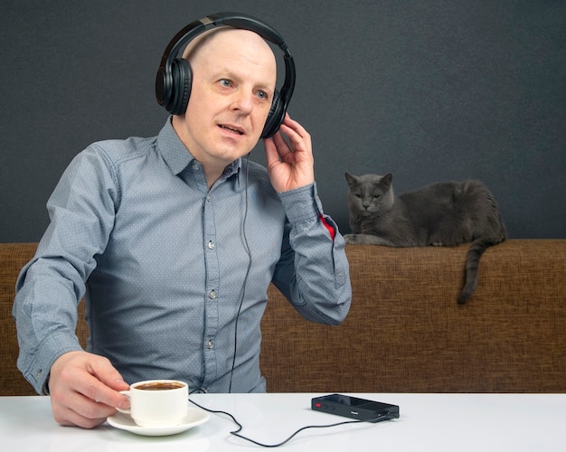 Homem com fones de ouvido, ouvindo música e tomando um café, sentado em um sofá com um gato cinza