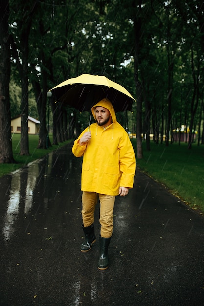 Homem com guarda-chuva caminhando no parque de verão em dia chuvoso. Homem com capa de chuva e botas de borracha, tempo chuvoso no beco