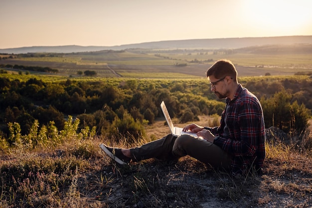 Homem com laptop sentado à beira de uma montanha com vistas deslumbrantes sobre o vale