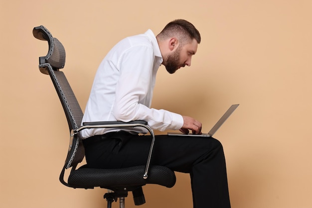 Homem com má postura sentado em uma cadeira e usando laptop contra um fundo laranja pálido