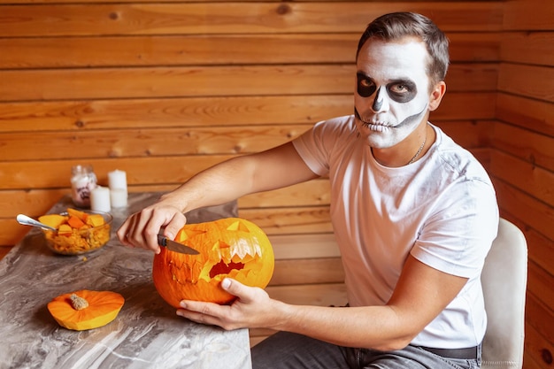 Homem com maquiagem artística assustadora se prepara para o Halloween esculpindo abóboras