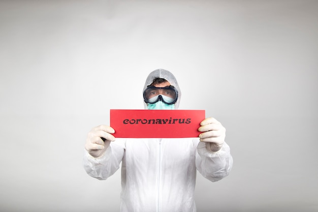 Homem com máscara cirúrgica, óculos, traje de proteção branco e letreiro vermelho isolado no estúdio em fundo branco. Nova epidemia de pandemia e novo coronavírus de rápida expansão.