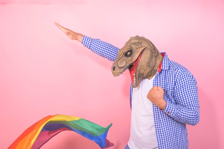 Foto homem com máscara de dinossauro com pose engraçada com uma bandeira de arco-íris em fundo rosa isolado