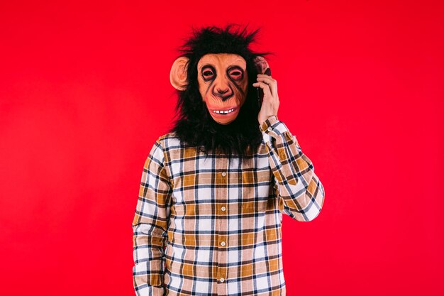 Homem com máscara de macaco chimpanzé e camisa quadriculada falando em seu celular ou smartphone em fundo vermelho