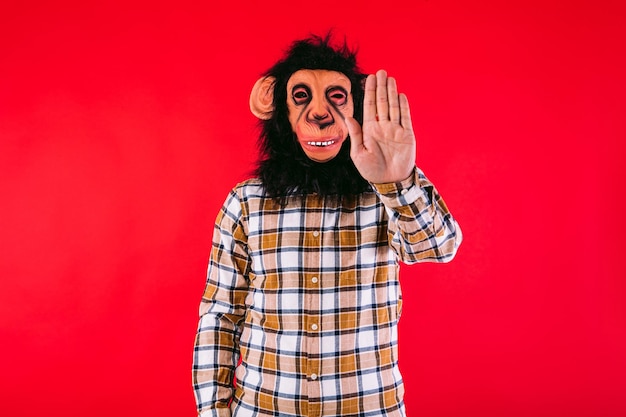 Homem com máscara de macaco chimpanzé e camisa quadriculada fazendo o sinal de stop com a mão no fundo vermelho
