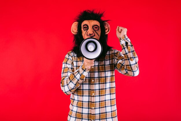 Homem com máscara de macaco chimpanzé e camisa xadrez gritando com um megafone em fundo vermelho