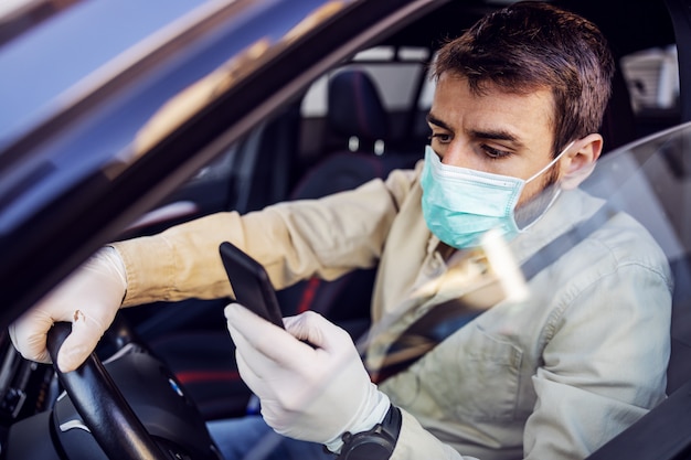 Homem com máscara e luvas dirigindo um carro digitando no celular smartphone. Prevenção de infecções e controle de epidemia. Pandemia mundial. Fique seguro.