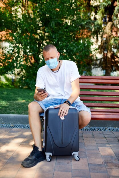 Homem com máscara protetora, sentado no banco do parque ao ar livre com uma mala e um telefone celular, vida durante a pandemia de coronavírus, viagem aérea de abertura, conceito de viagem.