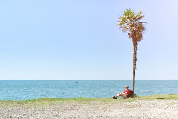 Homem com mochila senta-se sob uma palmeira alta no fundo do mar e céu azul