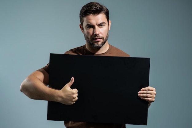 Homem com o polegar para cima segura banner no retrato de fundo cinza do estúdio de homem segurando cartaz em branco vazio m