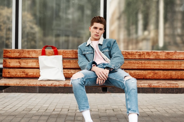 Homem com roupa de ganga azul e jeans sentado num banco de madeira